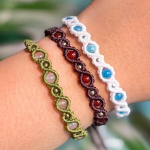 Op de afbeelding: Drie macramé armbanden in groen, bordeaux en wit, elk versierd met edelstenen. De groene armband heeft roze stenen, de bordeauxrode heeft rode stenen en de witte armband heeft blauwe stenen. De armbanden worden om een pols getoond.