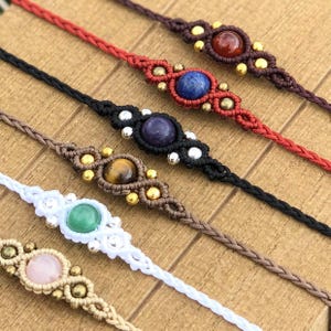 Puede incluir: Primer plano de varias pulseras de macramé. Cada pulsera presenta una piedra preciosa de diferente color, incluyendo rojo, azul, morado, marrón, verde y rosa. Las pulseras están hechas con cordones de varios colores y acentuadas con cuentas doradas y plateadas.