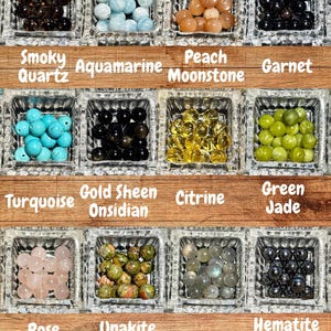 Op de afbeelding: Een verzameling edelsteen kralen in vierkante glazen bakjes. De kralen zijn in verschillende kleuren, waaronder rookkwarts, aquamarijn, perzikmaansteen, granaat, turkoois, goudglans obsidiaan, citrien, groene jade, rozenkwarts, unakiet, labradoriet en hematiet.
