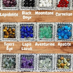 Op de afbeelding: Een verzameling gepolijste edelsteen kralen in kleine glazen bakjes. De afbeelding toont Lepidoliet, Zwarte Onyx, Maansteen, Carneool, Tijgeroog, Lapis Lazuli, Aventurijn, Apatiet, Mosagaat, Amethist, Prehniet en Ryoliet. De namen van de stenen staan onder elke bak.