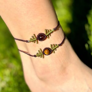 May include: Two macrame anklets with a dark brown cord and olive green accents. Each anklet features a central gemstone, one a dark red-brown, the other a golden-brown. The anklets are designed to be worn around the ankle.