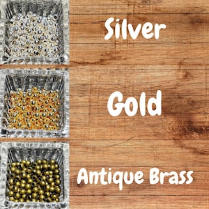 Op de afbeelding: Drie vierkante glazen bakjes gevuld met zilveren, gouden en antiek messingkleurige kralen. De woorden "Silver", "Gold" en "Antique Brass" staan in het wit op een houten achtergrond.