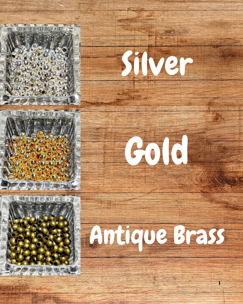 Op de afbeelding: Drie vierkante glazen bakjes gevuld met zilveren, gouden en antiek messingkleurige kralen. De woorden "Silver", "Gold" en "Antique Brass" staan in het wit op een houten achtergrond.
