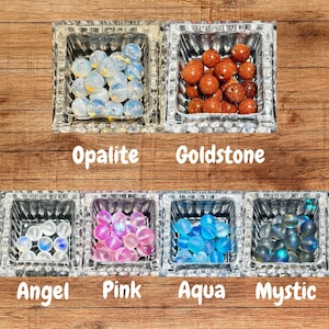 Op de afbeelding: Een assortiment kleurrijke, door de mens gemaakte kralen in vierkante glazen bakjes. De kralen zijn gelabeld met "Opalite", "Goldstone", "Angel", "Pink", "Aqua" en "Mystic". De tekst "Man Made" en "Matte Fiery Aura Quartz" zijn ook zichtbaar.