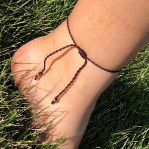 Op de afbeelding: Een bruine macram&eacute; enkelband met kleine goudkleurige kralen. De enkelband is om een enkel geknoopt en rust op groen gras. Het macram&eacute; koord is geknoopt om de maat aan te passen.