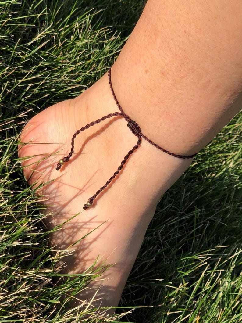 Op de afbeelding: Een bruine macram&eacute; enkelband met kleine goudkleurige kralen. De enkelband is om een enkel geknoopt en rust op groen gras. Het macram&eacute; koord is geknoopt om de maat aan te passen.