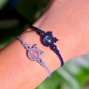 Könnte beinhalten: Zwei Makramee-Armbänder werden an einem Handgelenk gezeigt. Eines ist marineblau mit einem dunklen, irisierenden Stein. Das andere ist hellgrau mit einem rosafarbenen Stein. Beide Armbänder haben ein gewebtes Design mit dekorativen Knoten und Perlen.