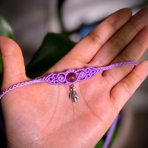 May include: A handmade macrame choker necklace in a soft lavender color. The necklace features a central amethyst stone and a small silver mushroom charm. The intricate knotwork and delicate design create a bohemian aesthetic.
