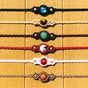 Op de afbeelding: Zes macram&eacute; armbanden met edelsteen-middelpunten worden getoond. De armbanden zijn zwart, beige, bruin, rood, wit en lichtbruin. Elke armband heeft een andere edelsteen, waaronder blauw, bruin, groen, wit, rood en oranje.