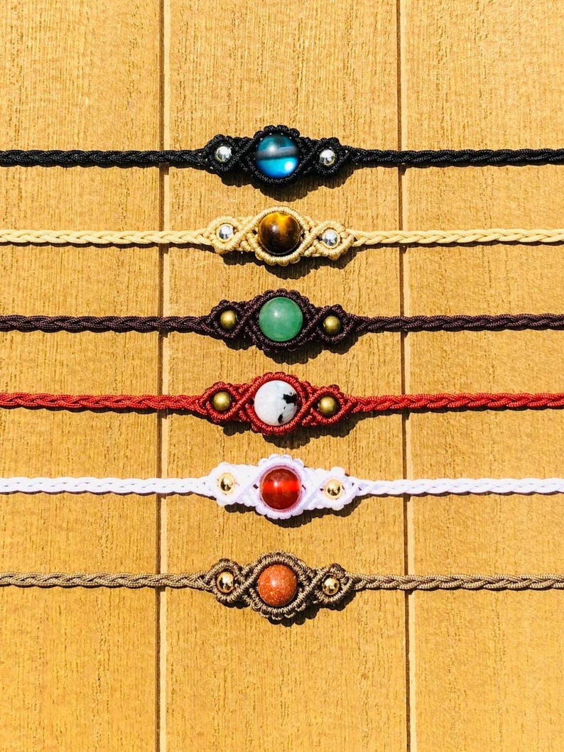 Op de afbeelding: Zes macram&eacute; armbanden met edelsteen-middelpunten worden getoond. De armbanden zijn zwart, beige, bruin, rood, wit en lichtbruin. Elke armband heeft een andere edelsteen, waaronder blauw, bruin, groen, wit, rood en oranje.