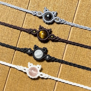 May include: Four macrame bracelets with gemstone details. The bracelets are in grey, brown, black, and white. Each bracelet has a different gemstone, including a black stone, tiger's eye, white stone, and a pink stone.