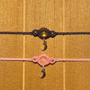 May include: Two macrame chokers. One is dark brown with a yellow orb and a silver crescent moon charm. The other is pink with a pink orb and a silver crescent moon charm. Both are on a wooden surface.