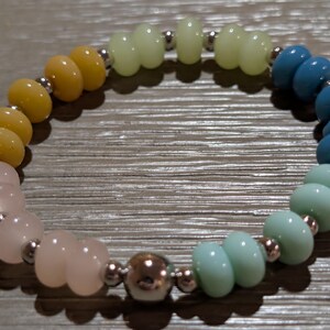 May include: A beaded bracelet featuring a rainbow of colors. The bracelet has a gradient of colors, starting with yellow, transitioning to pink, then green, and ending with blue. The beads are separated by small silver-colored beads.