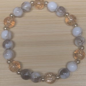 May include: A bracelet with a mix of white, marbled grey, and amber-coloured beads. The bracelet includes small silver-coloured spacer beads. The amber beads have a textured, faceted appearance, adding visual interest to the design.