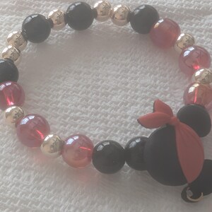 May include: A beaded bracelet featuring black, gold, and red beads. The bracelet includes a black charm with a red bow and a gold hoop earring. The beads are round and vary in size.