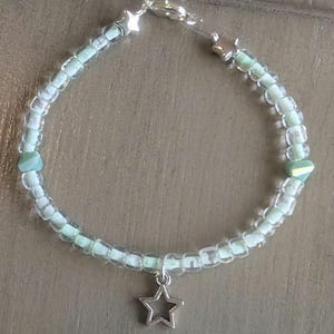 May include: A delicate beaded bracelet with clear and pale green beads. The bracelet has silver-toned star-shaped accents and a dangling star charm. It is secured with a silver clasp, creating a charming accessory.