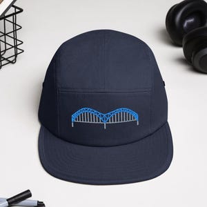 May include: Navy blue five-panel cap with a blue embroidered bridge design on the front. The cap has a flat brim and a structured crown. A stylish accessory for everyday wear.