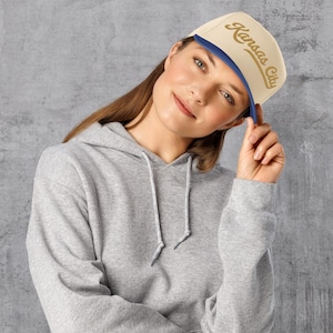 May include: A beige and blue baseball cap with the text "Kansas City" in gold script. The cap has a flat brim and a blue underside. The person in the image is wearing a gray hoodie.