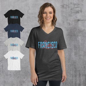 May include: A woman wearing a dark gray V-neck t-shirt with the word "FRANCISCO" in blue and red lettering. The image also shows four other t-shirts in black, navy, light gray, and white, each with the same design.