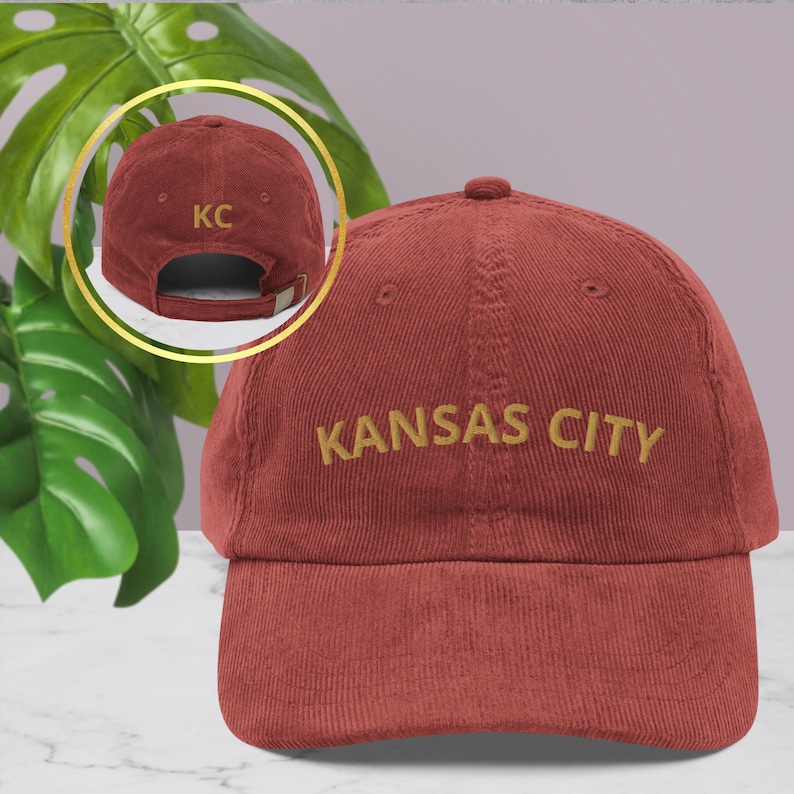 Puede incluir: Gorra de b&eacute;isbol de pana rojo ladrillo con "KANSAS CITY" bordado en hilo dorado en la parte delantera. La parte trasera de la gorra, que se muestra en un recuadro circular, presenta "KC" bordado en dorado. La gorra tiene una visera curva y una correa ajustable.