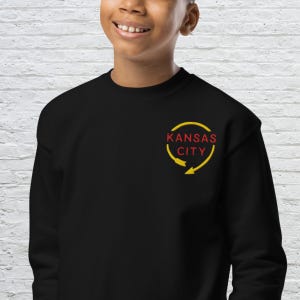 May include: Black sweatshirt with a yellow and red "Kansas City" logo. The logo features the city name in red, encircled by a yellow arrow design. The sweatshirt is a solid black colour.
