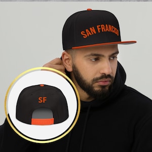 May include: Black flat-brimmed baseball cap with the text "SAN FRANCISCO" in orange. The cap has an orange brim and a matching orange adjustable snap closure. The back of the cap features the letters "SF" in orange.