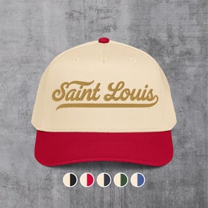 Puede incluir: Gorra de béisbol color crema con visera roja y botón rojo en la parte superior. El texto "Saint Louis" está bordado en oro en la parte delantera. Una fila de muestras de color en la parte inferior.
