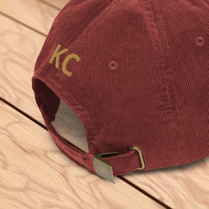 Puede incluir: Gorra de b&eacute;isbol de pana burdeos con las letras "KC" bordadas en dorado en el lateral. La gorra tiene un cierre de hebilla met&aacute;lica y est&aacute; sobre una superficie de madera.