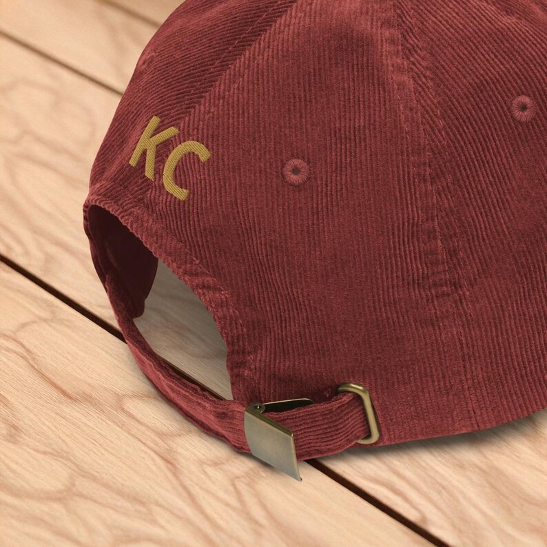 Puede incluir: Gorra de b&eacute;isbol de pana burdeos con las letras "KC" bordadas en dorado en el lateral. La gorra tiene un cierre de hebilla met&aacute;lica y est&aacute; sobre una superficie de madera.