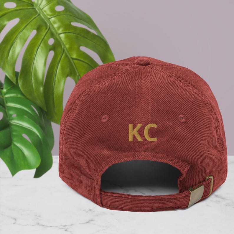 Puede incluir: Gorra de b&eacute;isbol de pana burdeos con las letras "KC" bordadas en dorado. La gorra tiene una visera curva y una correa ajustable con hebilla met&aacute;lica. El fondo incluye hojas verdes y una superficie de color p&uacute;rpura claro.