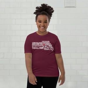 May include: A maroon t-shirt featuring a white graphic of the Kansas City outline with the text "Kansas City" and a red heart. The shirt is made of a soft material and is suitable for casual wear.