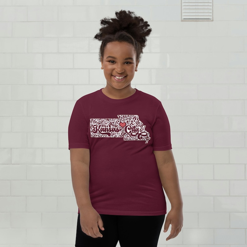 May include: A maroon t-shirt featuring a white graphic of the Kansas City outline with the text "Kansas City" and a red heart. The shirt is made of a soft material and is suitable for casual wear.