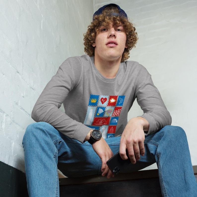 May include: A gray long-sleeve shirt featuring a colorful graphic of postage stamp designs. The stamps have various illustrations, including a crown, heart, and badminton shuttlecock. The shirt is paired with blue jeans and a dark watch.