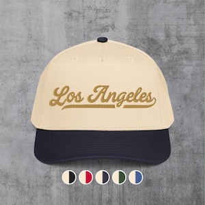 May include: A cream-colored baseball cap with a dark navy blue brim and a dark navy blue button on top. The text Los Angeles is embroidered in a gold-colored script font across the front panels. Five small circular color swatches are below.