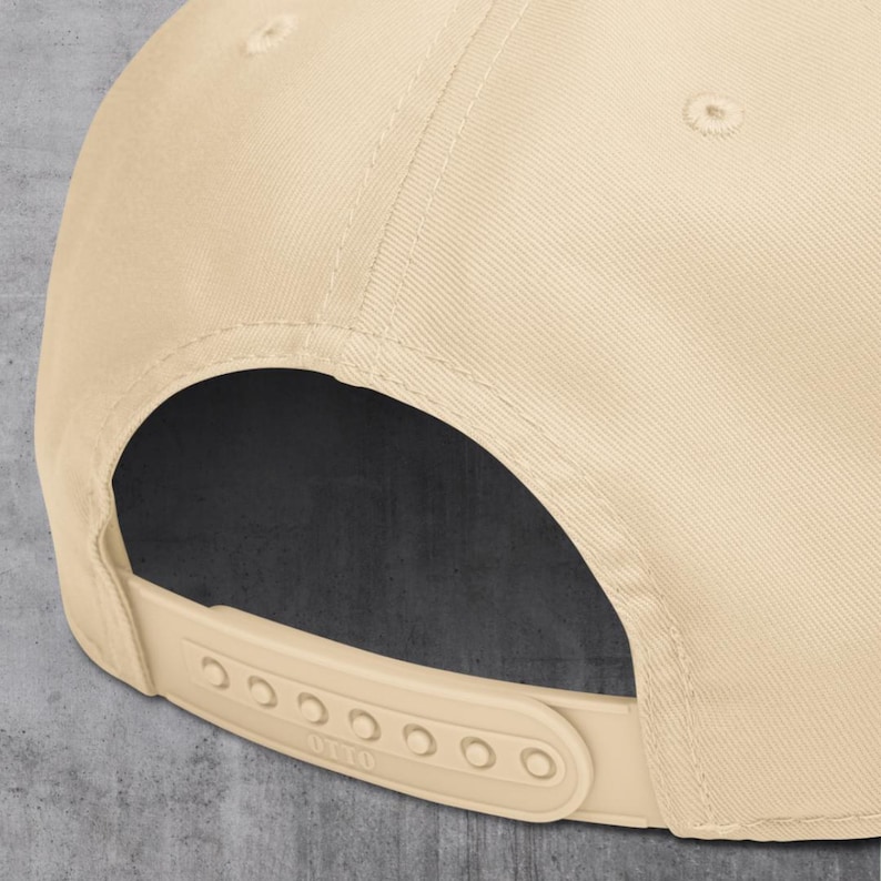May include: A beige baseball cap with a flat brim and a snapback closure. The cap is made of a textured fabric and features a curved visor. The adjustable strap has multiple holes for sizing.