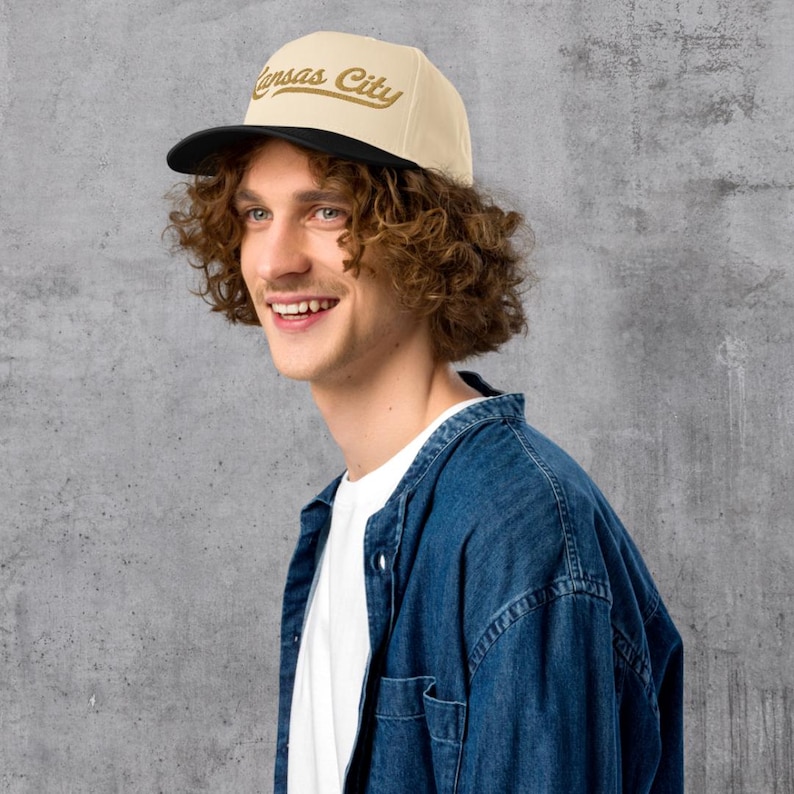 May include: A beige and black flat-brimmed cap with the words "Kansas City" in gold script. The cap has a black brim and a beige crown. The person in the image is wearing a denim shirt.