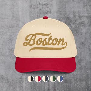 May include: A baseball cap with a cream-colored crown and a red brim. The word "Boston" is embroidered in a gold script across the front. The cap has a red button on top.