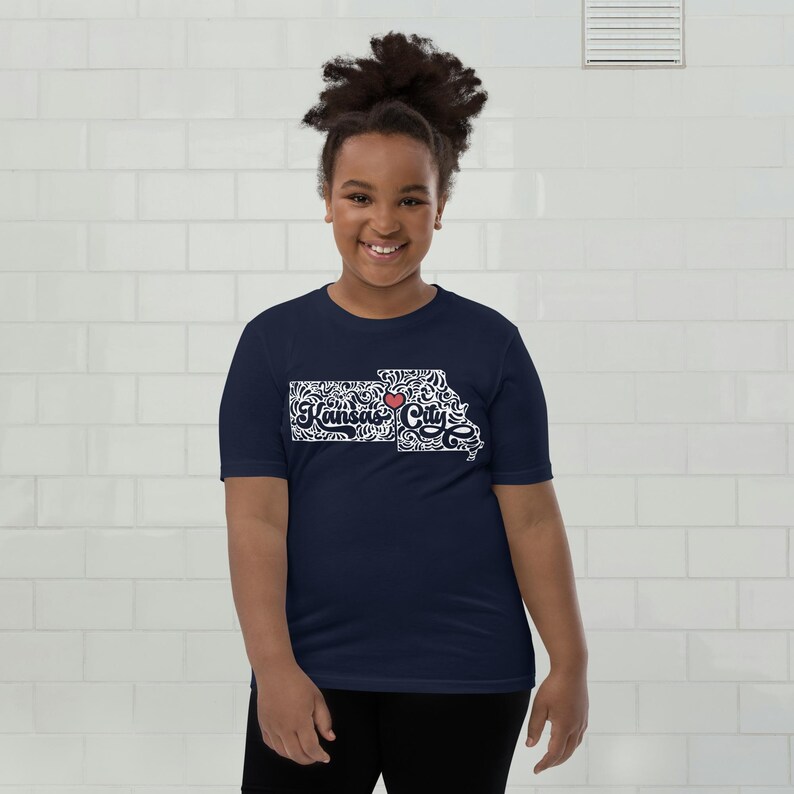 May include: A navy blue t-shirt featuring a white and black design of the Kansas state outline with the words "Kansas City" and a red heart. The shirt is worn by a person.