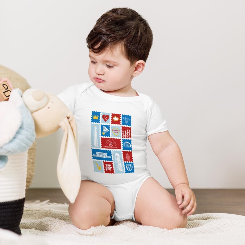 May include: White baby bodysuit with a colorful graphic of Kansas City landmarks and the text "Kansas City" in a stamp-like design. The design includes red, blue, and yellow elements. The baby is sitting on a white blanket.