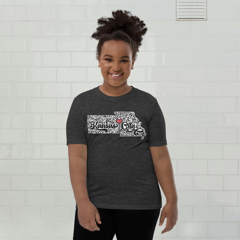May include: A dark gray t-shirt featuring a white and black design of the Kansas City outline with the text "Kansas City" and a red heart. The shirt is short-sleeved and made of a soft material.