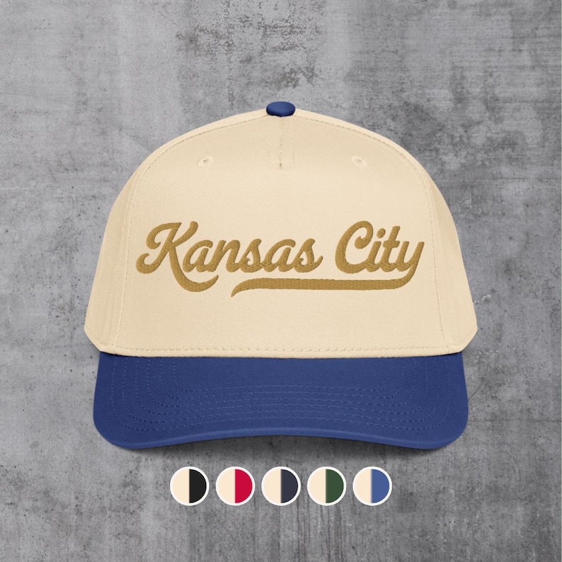 May include: A baseball cap with a cream-colored crown and a blue brim. The text "Kansas City" is embroidered in a gold script across the front. The cap has a blue button on top.