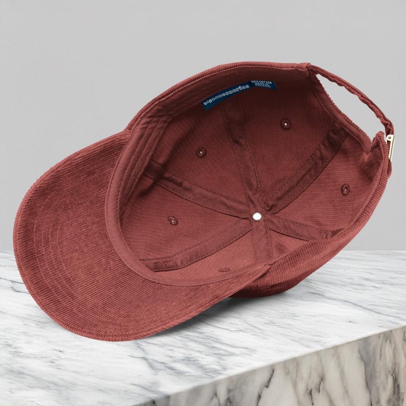Puede incluir: Gorra de b&eacute;isbol de pana granate, vista desde el interior, mostrando las costuras y la construcci&oacute;n. La gorra tiene una visera curva y una correa ajustable con una hebilla dorada. El interior presenta una etiqueta con texto.