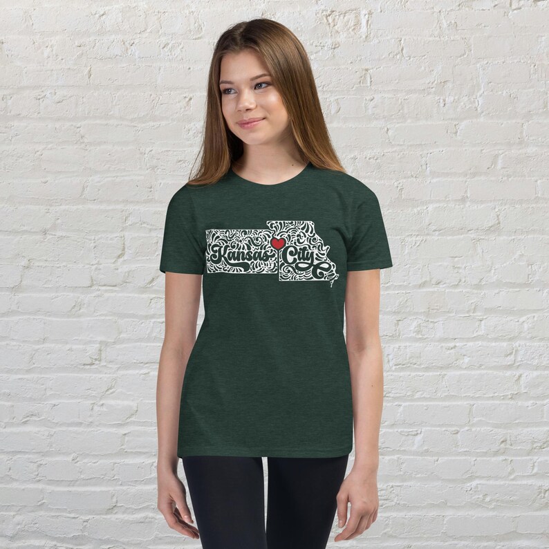 May include: Dark green t-shirt featuring a white outline of the state of Kansas with the words "Kansas City" in a decorative font. A red heart is placed over the city. The shirt is made of a soft material.