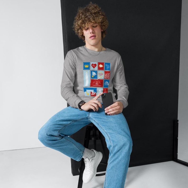 May include: A gray long-sleeve shirt featuring a colorful stamp design with various images and the text "KANSAS CITY". The shirt is made of a soft material and has a crew neck.