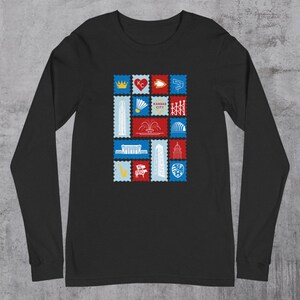 May include: Black long-sleeve shirt featuring a colorful stamp design with various Kansas City icons. The stamps include a crown, heart, arrow, and the words "Kansas City". The shirt is made of a soft material.