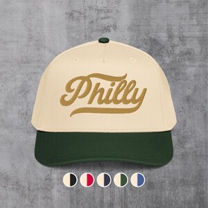 Puede incluir: Gorra de béisbol con corona color crema y visera verde oscuro. La palabra "Philly" está bordada en un guion dorado en la parte delantera. La gorra tiene un botón verde oscuro en la parte superior.