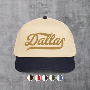 May include: A baseball cap with a cream-colored crown and a navy blue brim. The word "Dallas" is embroidered in a gold script across the front. The cap has a dark blue button on top.