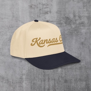 May include: A baseball cap with a cream-colored crown and a navy blue brim. The script "Kansas C" is embroidered in a gold color on the front of the cap. The cap has a curved brim and a small black button on top.