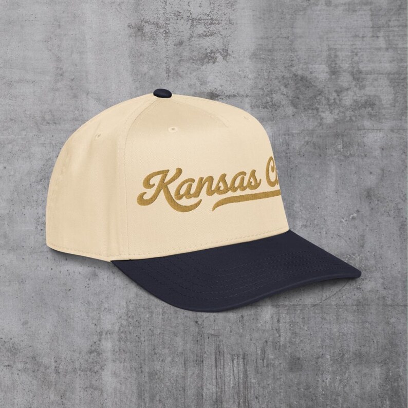 May include: A baseball cap with a cream-colored crown and a navy blue brim. The script "Kansas C" is embroidered in a gold color on the front of the cap. The cap has a curved brim and a small black button on top.