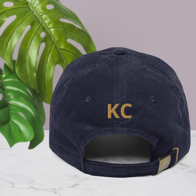 Puede incluir: Gorra de b&eacute;isbol de pana azul marino con las letras "KC" bordadas en dorado. La gorra tiene una visera curva y una correa ajustable con una hebilla de metal dorado. En el fondo, una gran hoja verde.